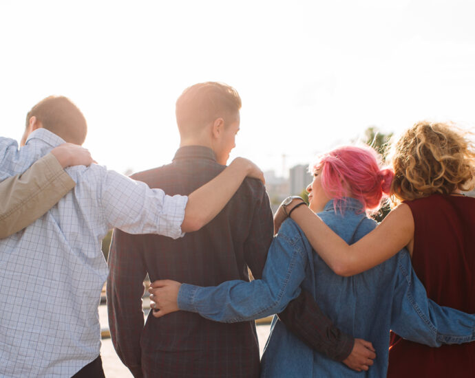 Back view of a diverse group of 8 young people, with their arms around the people on each side of them. Unity and support. Friendship together.