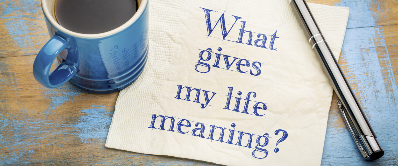 A handwritten sketched note, which reads, "What gives my life meaning?" written in blue ink on a white paper napkin, with a cup of espresso coffee and a silver pen, sitting on a worn, blue-painted wooden table.