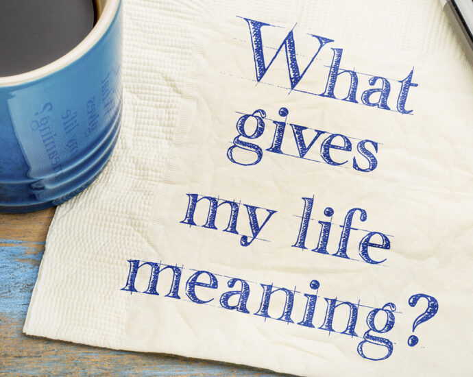 A handwritten sketched note, which reads, "What gives my life meaning?" written in blue ink on a white paper napkin, with a cup of espresso coffee and a silver pen, sitting on a worn, blue-painted wooden table.