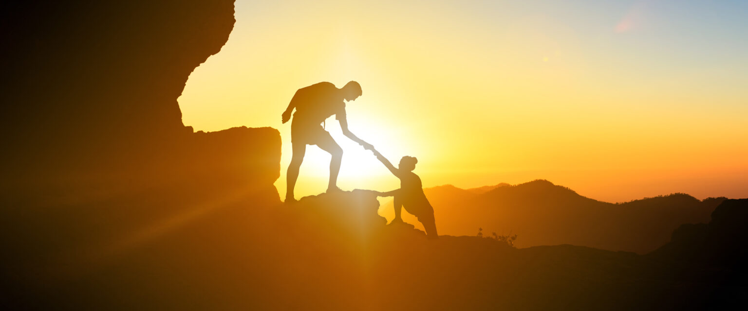 A photo with the silhouette of a man and a woman, as he gives her a helping hand, while climbing up a mountain. The image, taken in the mountains, shows a beautiful, golden sunset on Gran Canaria