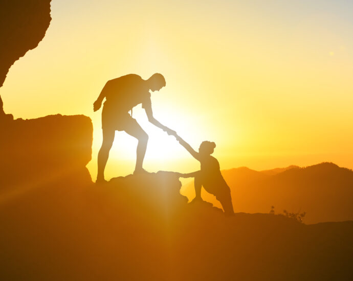 A photo with the silhouette of a man and a woman, as he gives her a helping hand, while climbing up a mountain. The image, taken in the mountains, shows a beautiful, golden sunset on Gran Canaria