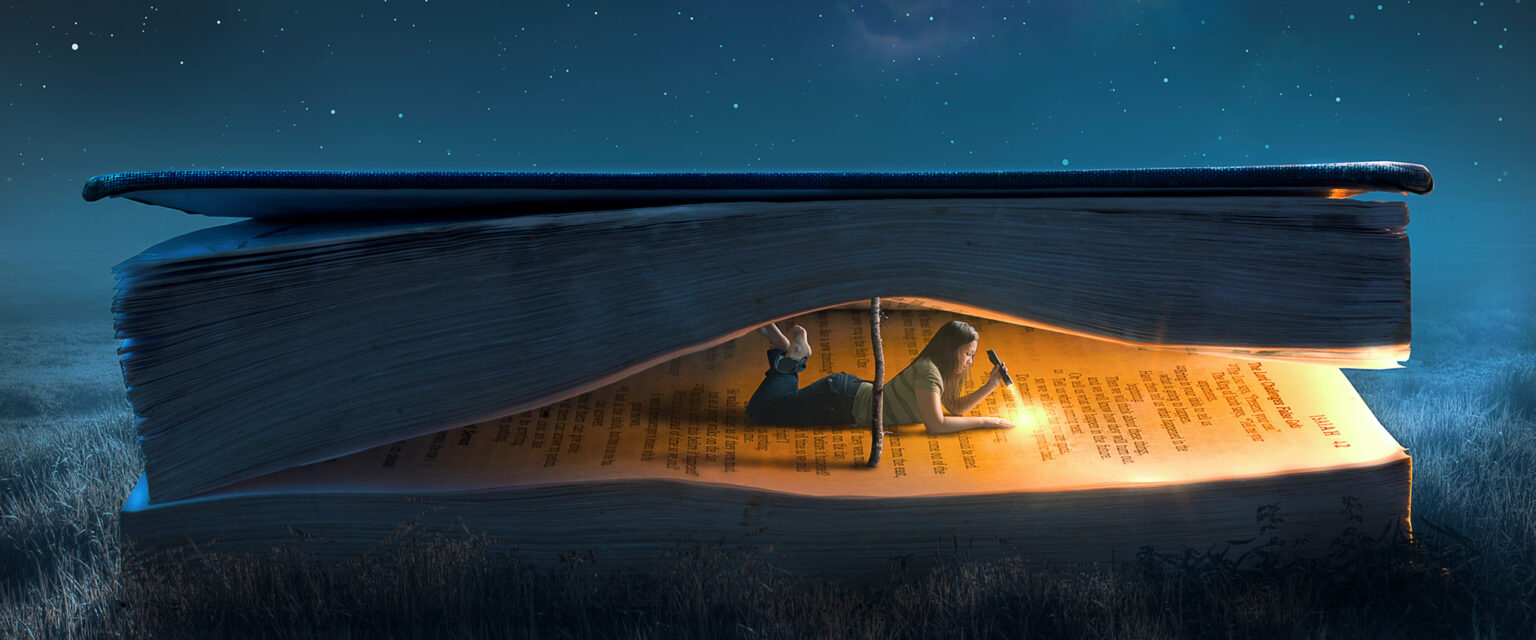 A young woman lies inside a large book, using a branch to prop it open, and reads by flashlight under a night sky.