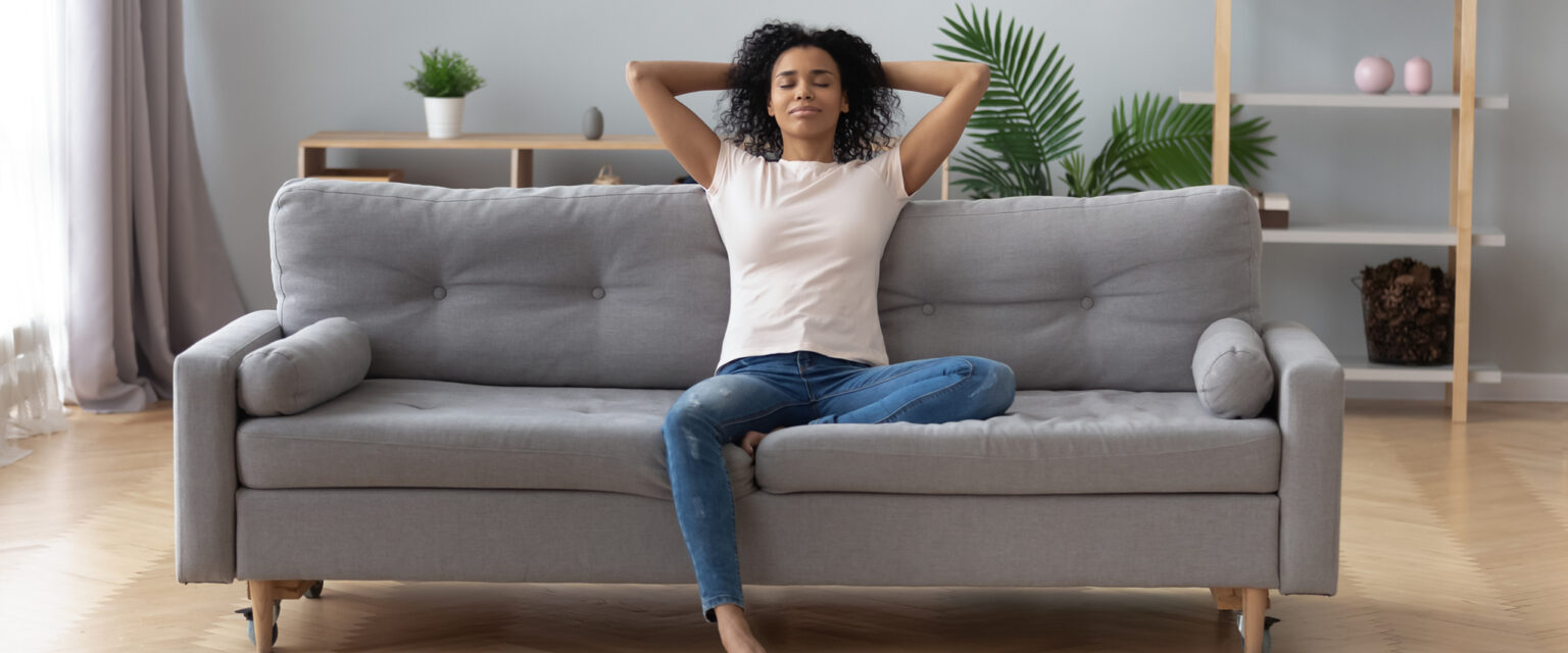 A young black woman sits on a gray sofa in a modern living room. She appears peaceful and stress-free while relaxing at home.
