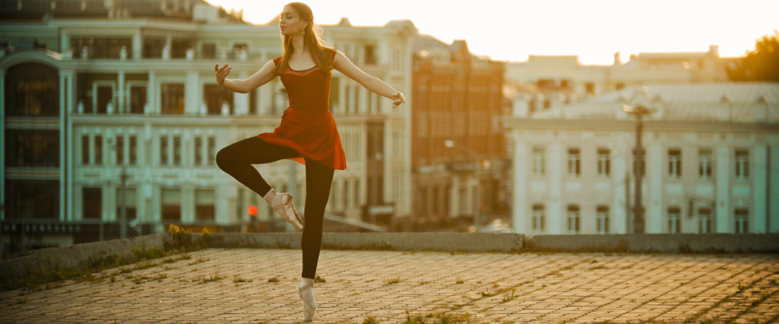 A young, inspired ballerina wearing a red dress, black tights, and gold ballet slippers, stands in a graceful pose atop a roof in an established old-world city.