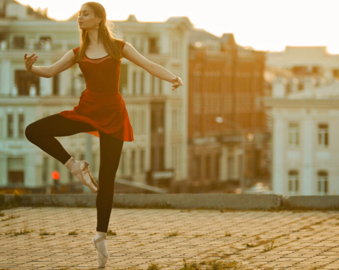 A young, inspired ballerina wearing a red dress, black tights, and gold ballet slippers, stands in a graceful pose atop a roof in an established old-world city.