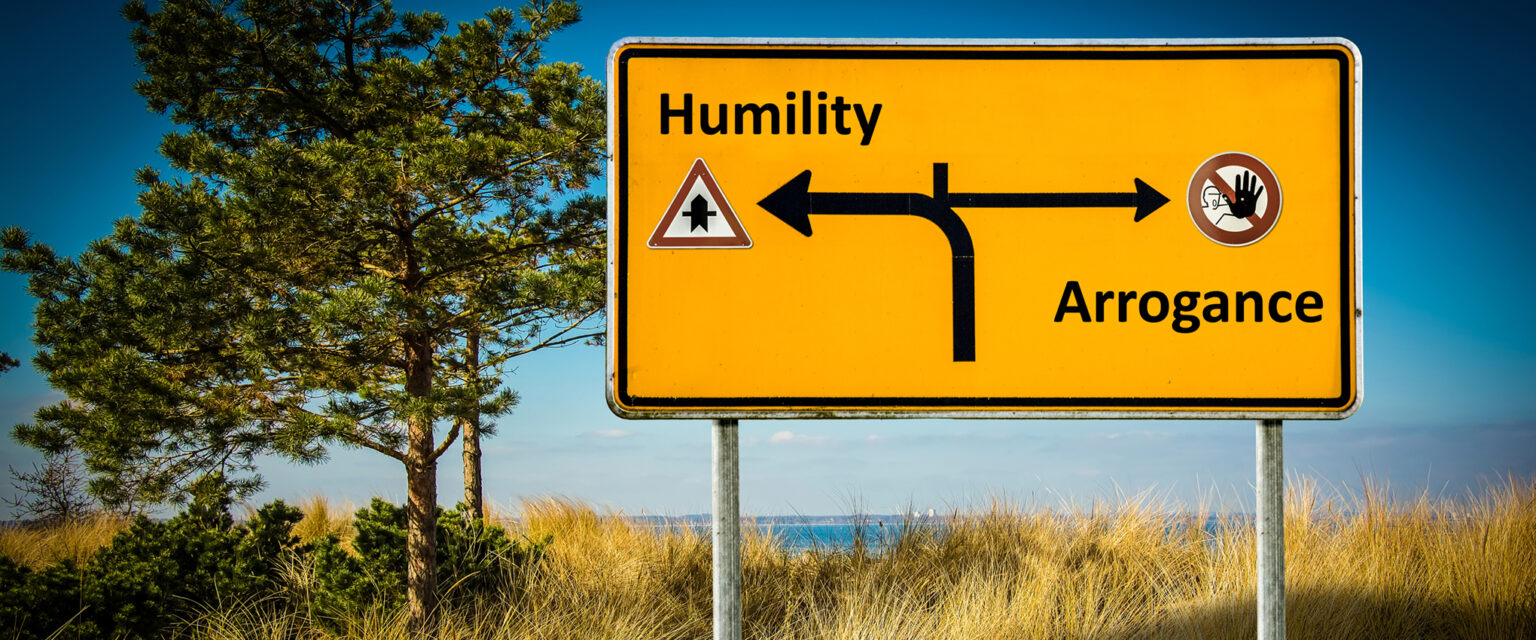 A yellow and black road sign, it shows a road curving to the left toward Humility and a caution symbol in the direction of Arrogance.