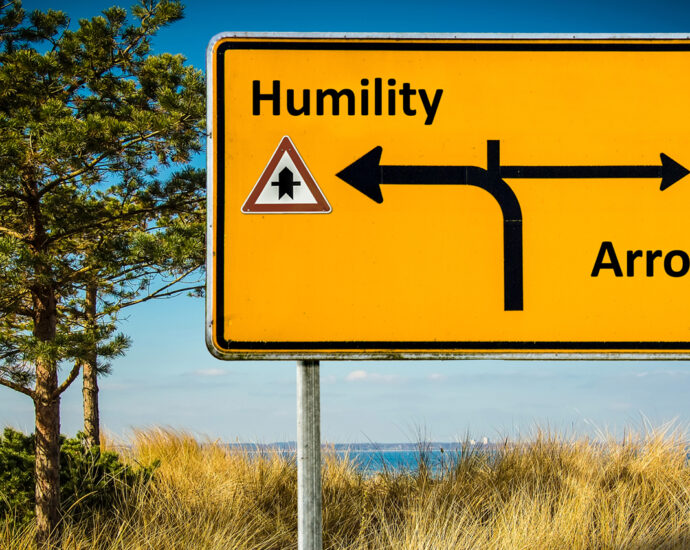 A yellow and black road sign, it shows a road curving to the left toward Humility and a caution symbol in the direction of Arrogance.