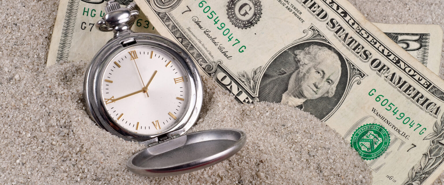 A photo of six dollars and a silver pocket watch, with a white face and gold Roman numerals, sitting in sand. The image represents lost time and money.