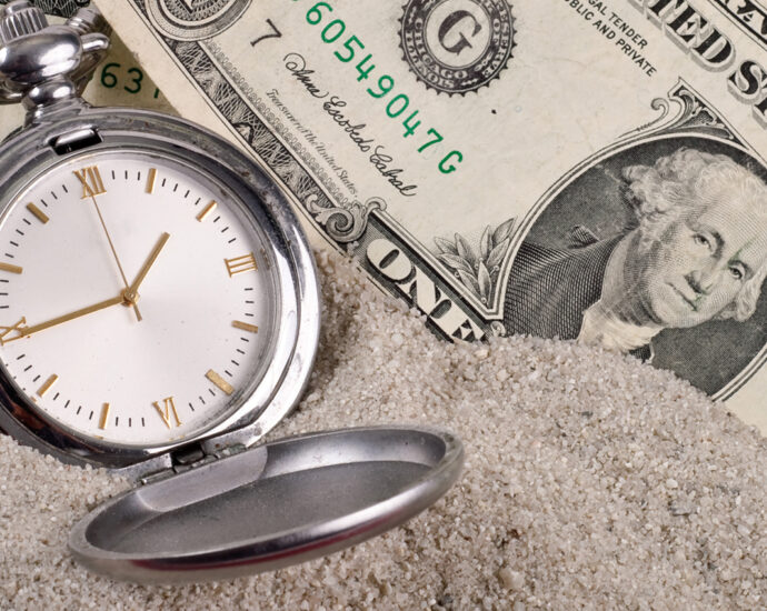 A photo of six dollars and a silver pocket watch, with a white face and gold Roman numerals, sitting in sand. The image represents lost time and money.