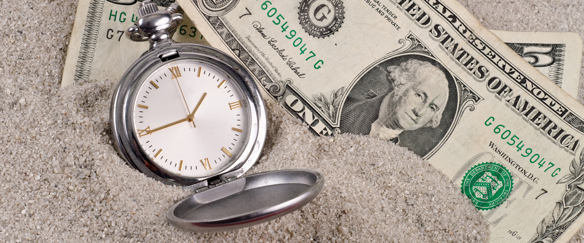 A photo of six dollars and a silver pocket watch, with a white face and gold Roman numerals, sitting in sand. The image represents lost time and money.