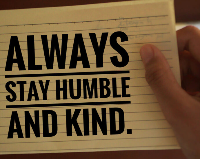 A young woman holds in her hand a piece of yellow, lined, spiral-bound notepad paper with the words "Always stay humble and kind" printed on it in black, block letters. Photographed on a medium brown background.