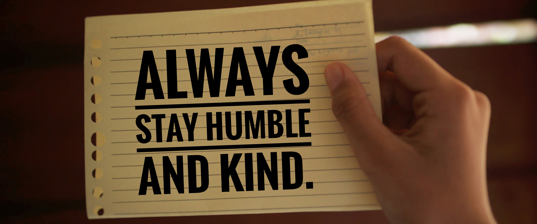 A young woman holds in her hand a piece of yellow, lined, spiral-bound notepad paper with the words "Always stay humble and kind" printed on it in black, block letters. Photographed on a medium brown background.