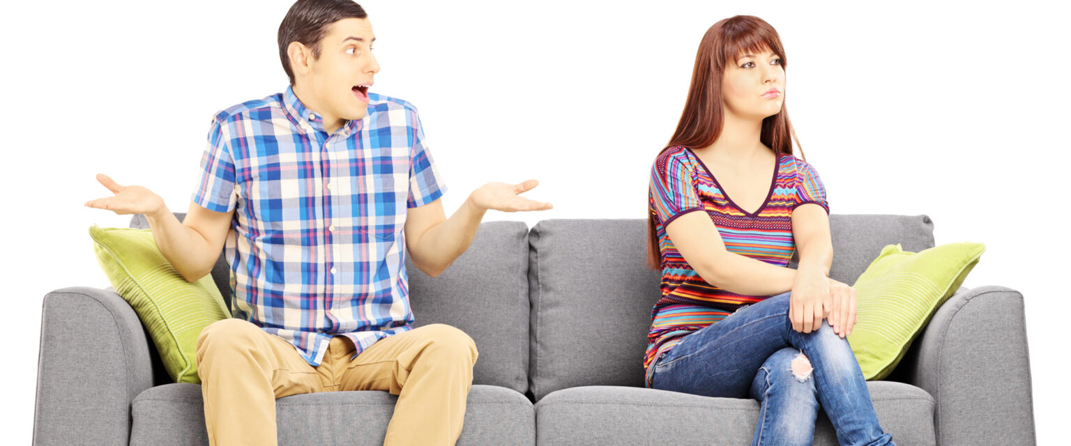 A young couple is sitting on a gray couch with green pillows, during an argument. The woman is looking away, with a stern expression on her face. The man is looking at her with his mouth agape, eyes wide, arms bent at the elbow with palms up, as if to say, "What did I say?!" This image is isolated on white background