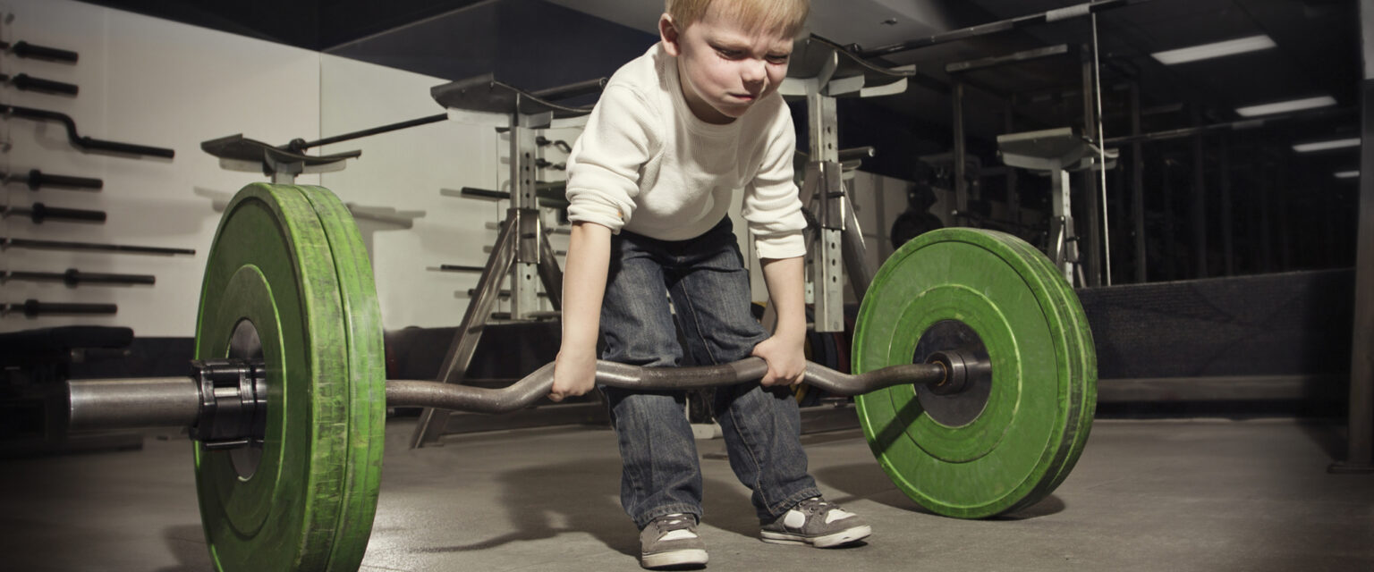 A determined little blonde-haired boy is trying to lift a barbell with large green weights.