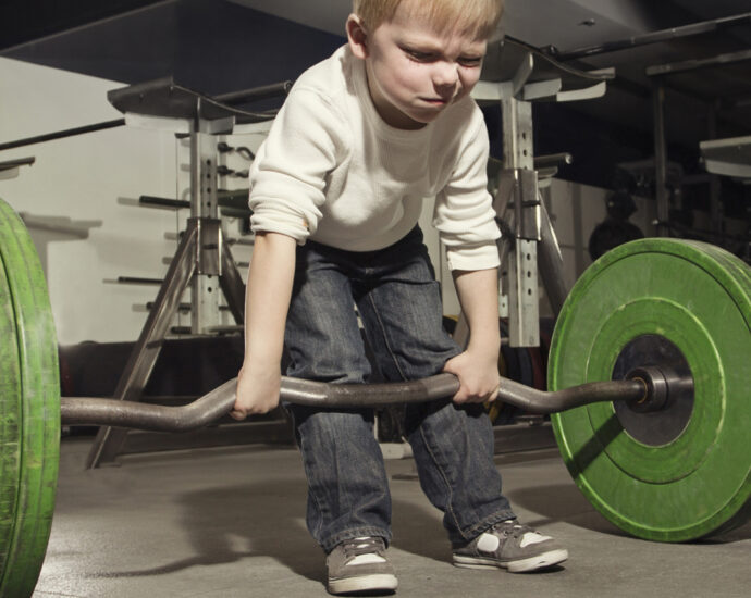 A determined little blonde-haired boy is trying to lift a barbell with large green weights.