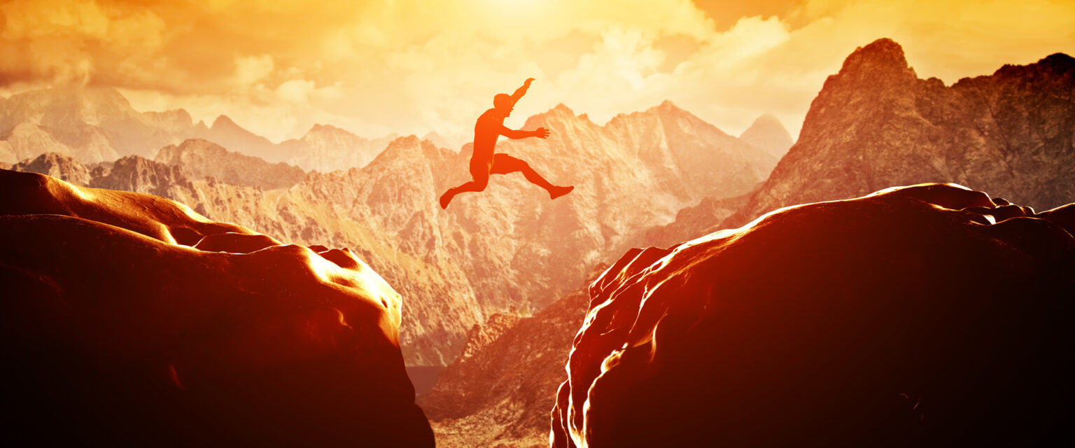 A man jumps over a divide between two rocky mountains at sunset. Freedom, risk, challenge, success.