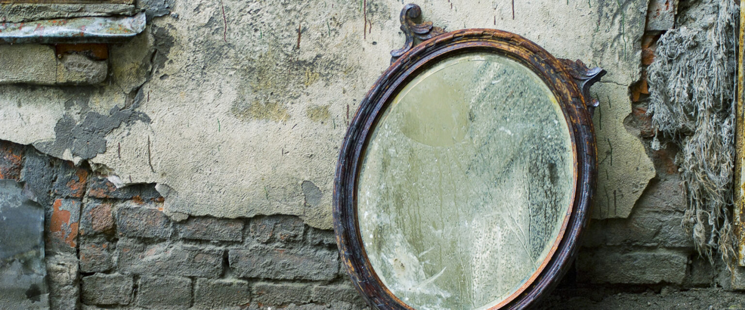 A photo of an old, wood-framed, Victorian era mirror with one of its two flourishes broken off. It is leaning against a worn and cracked wall with exposed brick at the bottom and up the side. The heavily worn and marked wooden frame, dirty glass, and damaged silver backing decry the mirror's age.