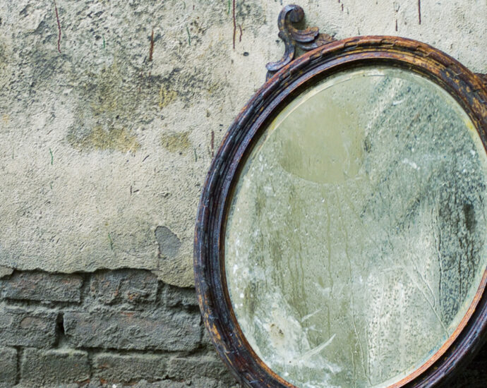 A photo of an old, wood-framed, Victorian era mirror with one of its two flourishes broken off. It is leaning against a worn and cracked wall with exposed brick at the bottom and up the side. The heavily worn and marked wooden frame, dirty glass, and damaged silver backing decry the mirror's age.