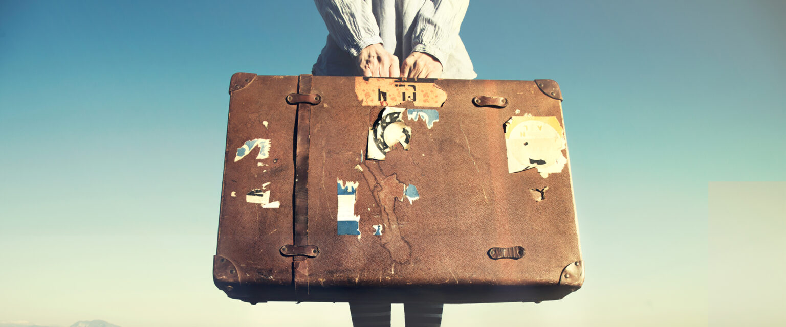 A woman stands on a long, wooden dock that stretches out into the distance. She is wearing a lightweight, ivory-colored jacket, striped stockings, and a pair of Converse Chuck Taylor, All Star, high-top sneakers. With both hands, she holds an old suitcase in front of her. It is made of light brown leather, is water-stained, and has a handful of torn and worn out pieces of travel stickers stuck to its front. One of the matching, leather securing straps is missing.