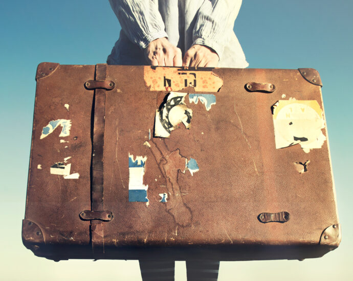 A woman stands on a long, wooden dock that stretches out into the distance. She is wearing a lightweight, ivory-colored jacket, striped stockings, and a pair of Converse Chuck Taylor, All Star, high-top sneakers. With both hands, she holds an old suitcase in front of her. It is made of light brown leather, is water-stained, and has a handful of torn and worn out pieces of travel stickers stuck to its front. One of the matching, leather securing straps is missing.