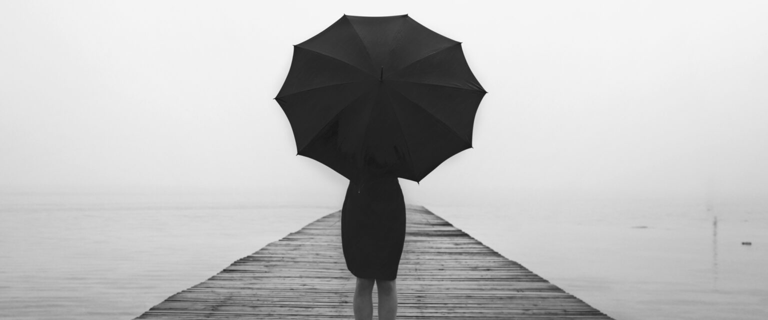 A black and white photo of an elegant woman, dressed in a black, knee-length skirt, and with an open black umbrella. She is standing with her back to the camera, on a wood-planked dock that fades into the foggy distance.