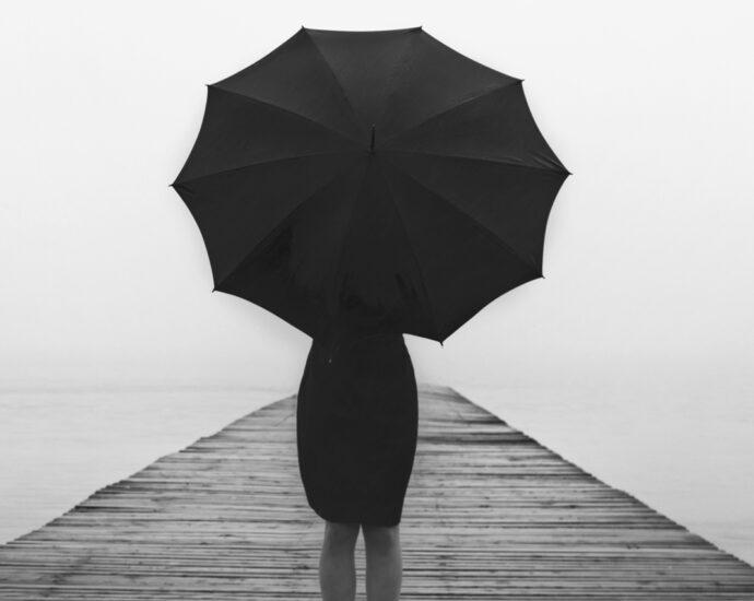 A black and white photo of an elegant woman, dressed in a black, knee-length skirt, and with an open black umbrella. She is standing with her back to the camera, on a wood-planked dock that fades into the foggy distance.