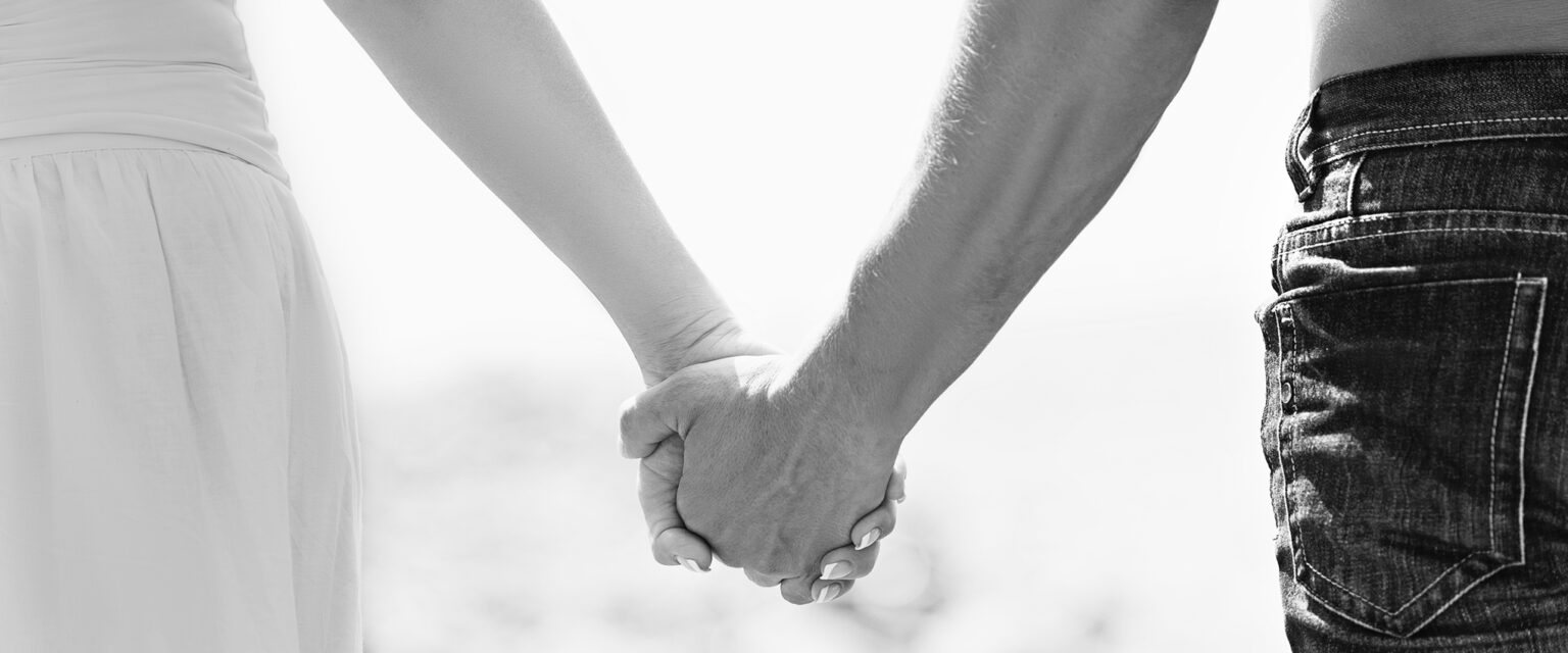 A black and white close-up photo of a woman, in a white skirt and top, and a man, wearing jeans and no shirt, holding hands as they walk together.