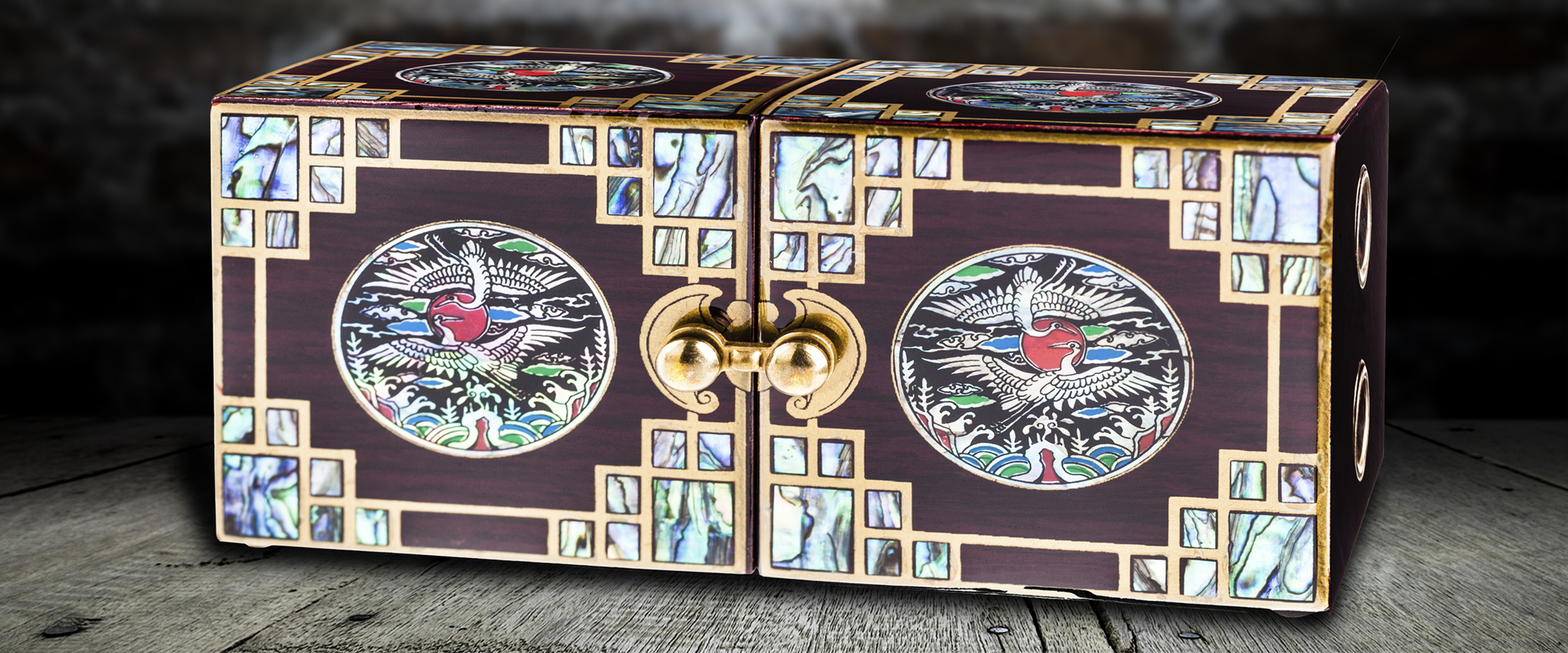 A luxurious Chinese wood and brass jewelry box, with abalone inlay and a bird design in the center of each panel. The box is sitting on a weathered wooden table with an old dark brick wall in the background