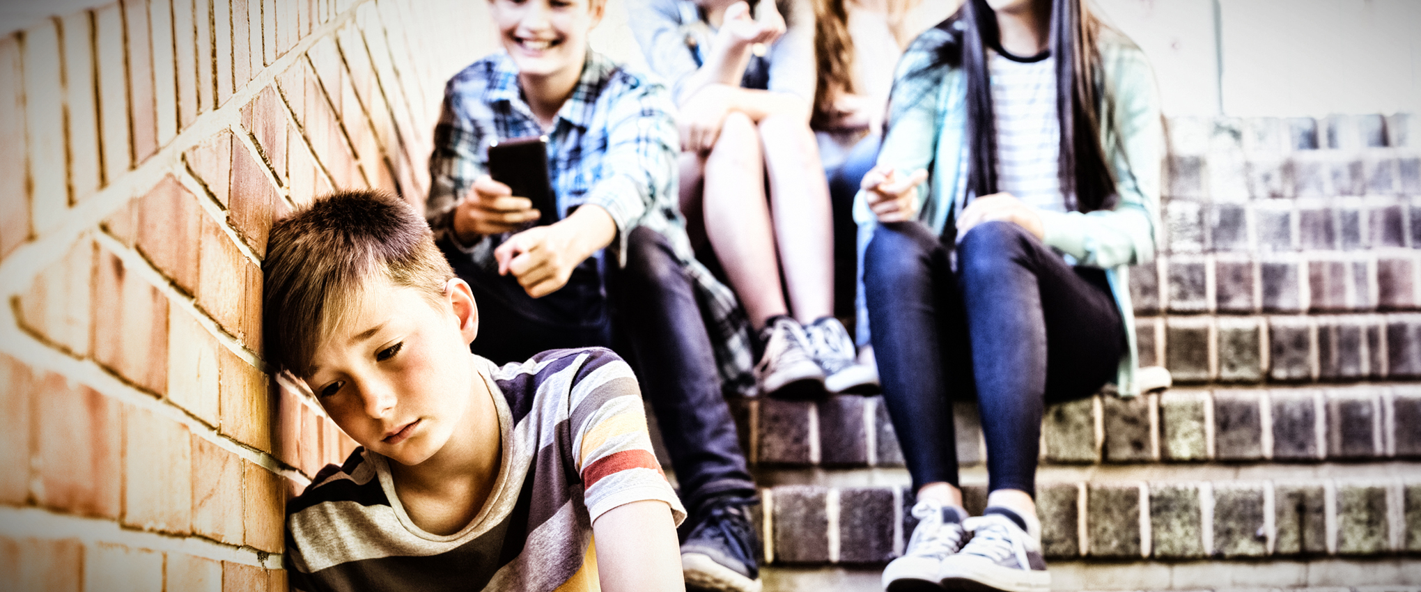 School kid bullies sit on a brick stairway above a boy who is seated, with his head against a brick wall. The victim's expression shows feelings of rejection and ridicule.