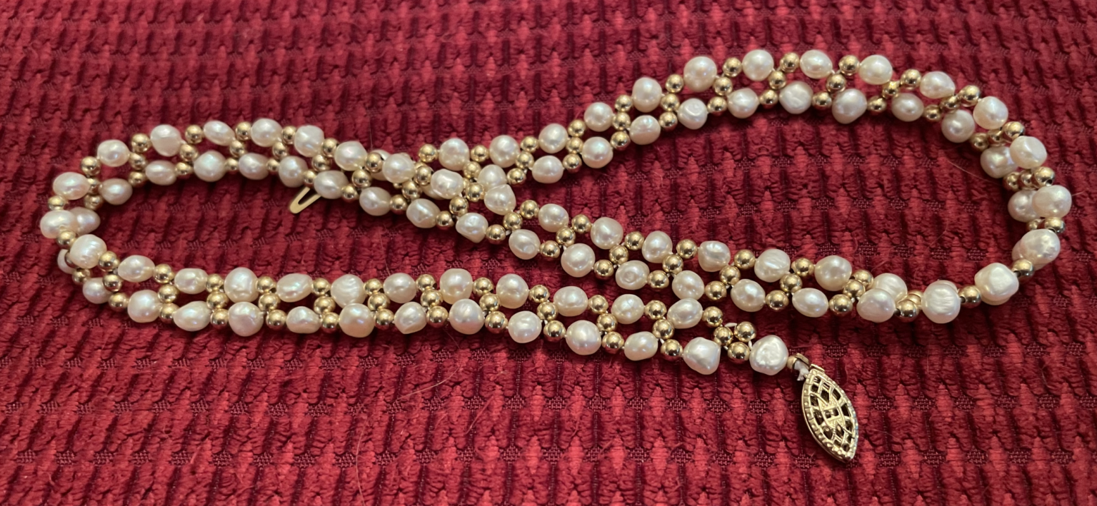 A beautiful double string of imperfect pearls with three small, gol beads between each pair of pearls. The necklace is laid out in the shape of the infinity symbol. They sit atop a heavy red fabric with a pattern made of dark triangles and light diamonds.