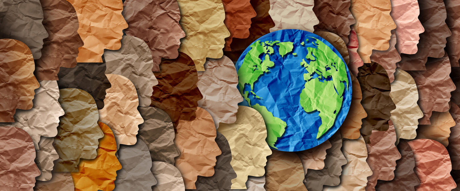 Paper cut-outs of people's heads and the Earth. Each of the items is wrinkled and overlaps those to the right of it. Each of the faces is cut from paper, which is reflective of the diverse skin colors of the human race. The Earth is cut from blue and green papers. All of the faces are pointed to the right, showing the concept of working together for progress.