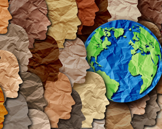 Paper cut-outs of people's heads and the Earth. Each of the items is wrinkled and overlaps those to the right of it. Each of the faces is cut from paper, which is reflective of the diverse skin colors of the human race. The Earth is cut from blue and green papers. All of the faces are pointed to the right, showing the concept of working together for progress.