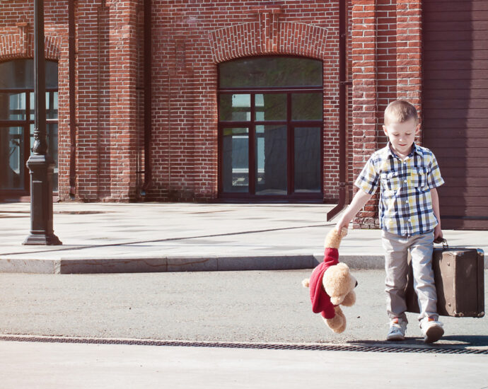 A little kid, carrying a vintage suitcase and teddy bear starts to cross the street on a bright sunny Summer morning.