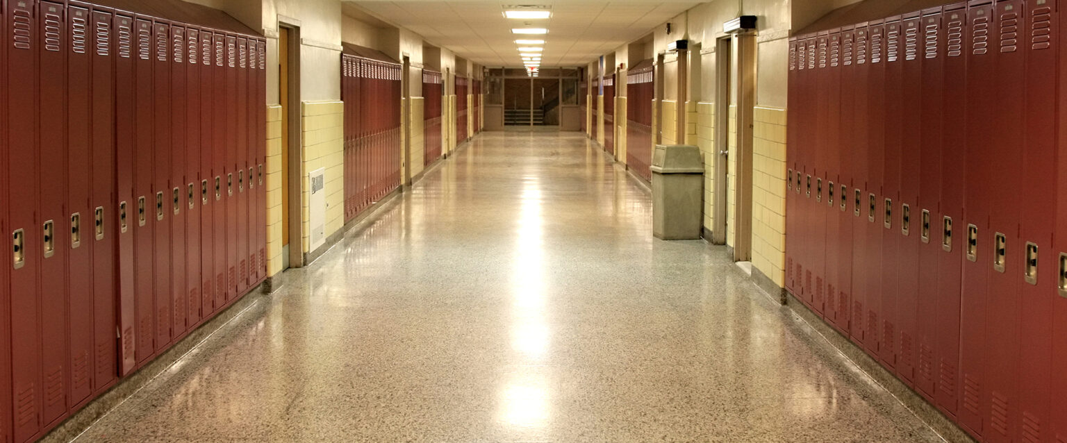 An empty school hallway with rust-colored, painted student lockers and yellow-beige walls. A single tan, covered garbage can sits aside one of the walls.