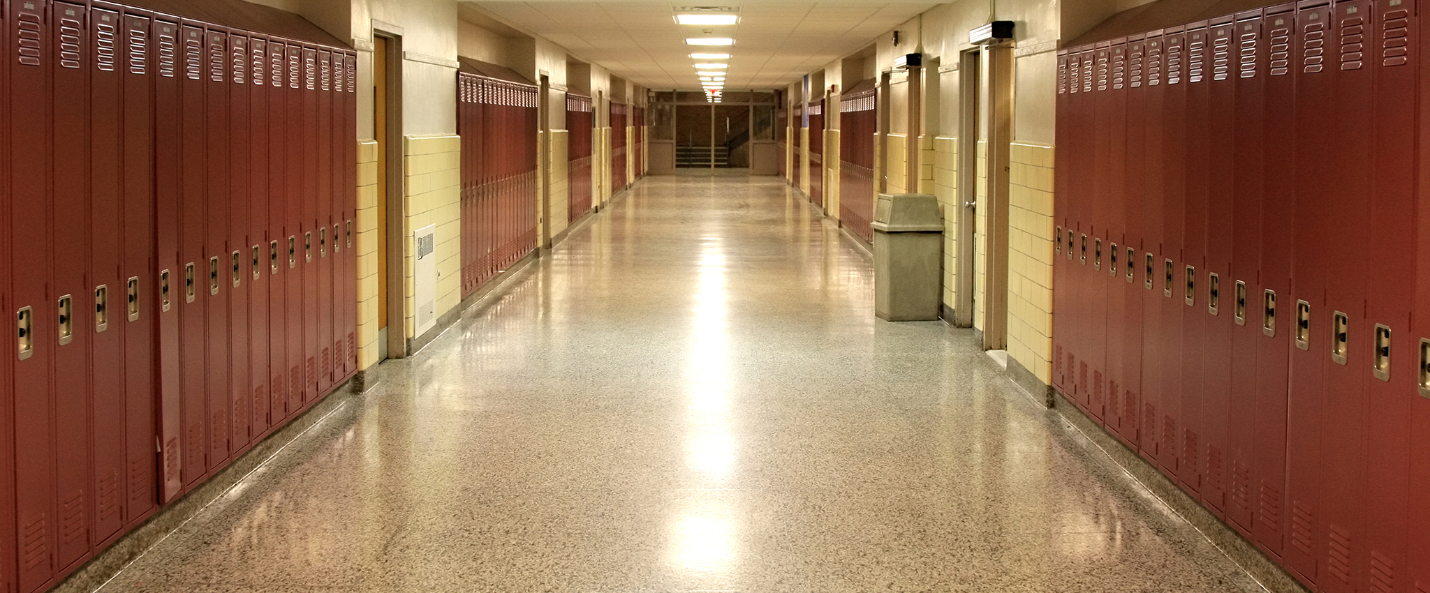 An empty school hallway with rust-colored, painted student lockers and yellow-beige walls. A single tan, covered garbage can sits aside one of the walls.