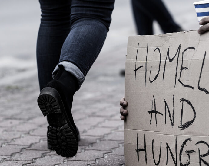A bearded young man with long hair, tan pants and lined coat, sits on a brick-paved walkway, holding a blue and white striped paper cup, and a cardboard sign with the inscription 'Homeless and hungry.' People in winter clothing walk past.
