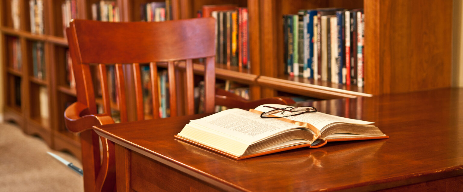 Library reading room. Empty wooden swivel chair at wooden library table with book open on the table. Glasses on top of book. Wooden bookcases with blurred bindings of colorful books behind table and chair. A steak knife lies on the floor nearby.