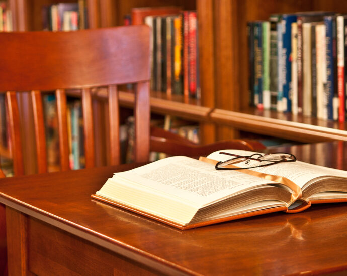 Library reading room. Empty wooden swivel chair at wooden library table with book open on the table. Glasses on top of book. Wooden bookcases with blurred bindings of colorful books behind table and chair. A steak knife lies on the floor nearby.