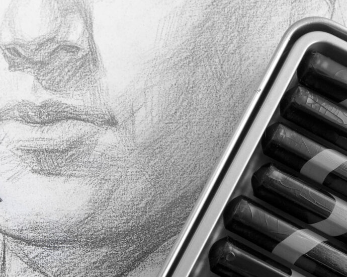 A pencil sketch of a man's face. A tin of numbered, black sketching pencils and a single red pencil lay across the art.