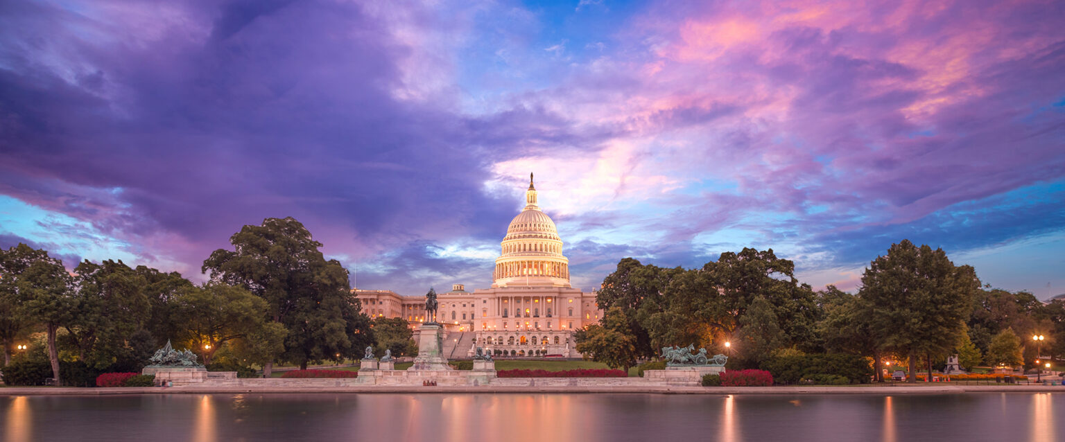 The US Capitol building dome is aglow as the sun sets for the evening. The blue sky is partially covered by purple and pink clouds colored by the setting sun. Shot from the water across from the halls of Congress.