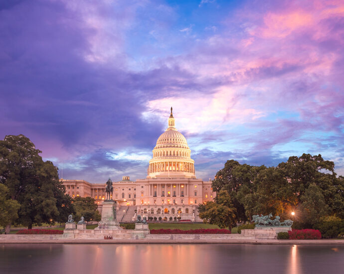 The US Capitol building dome is aglow as the sun sets for the evening. The blue sky is partially covered by purple and pink clouds colored by the setting sun. Shot from the water across from the halls of Congress.