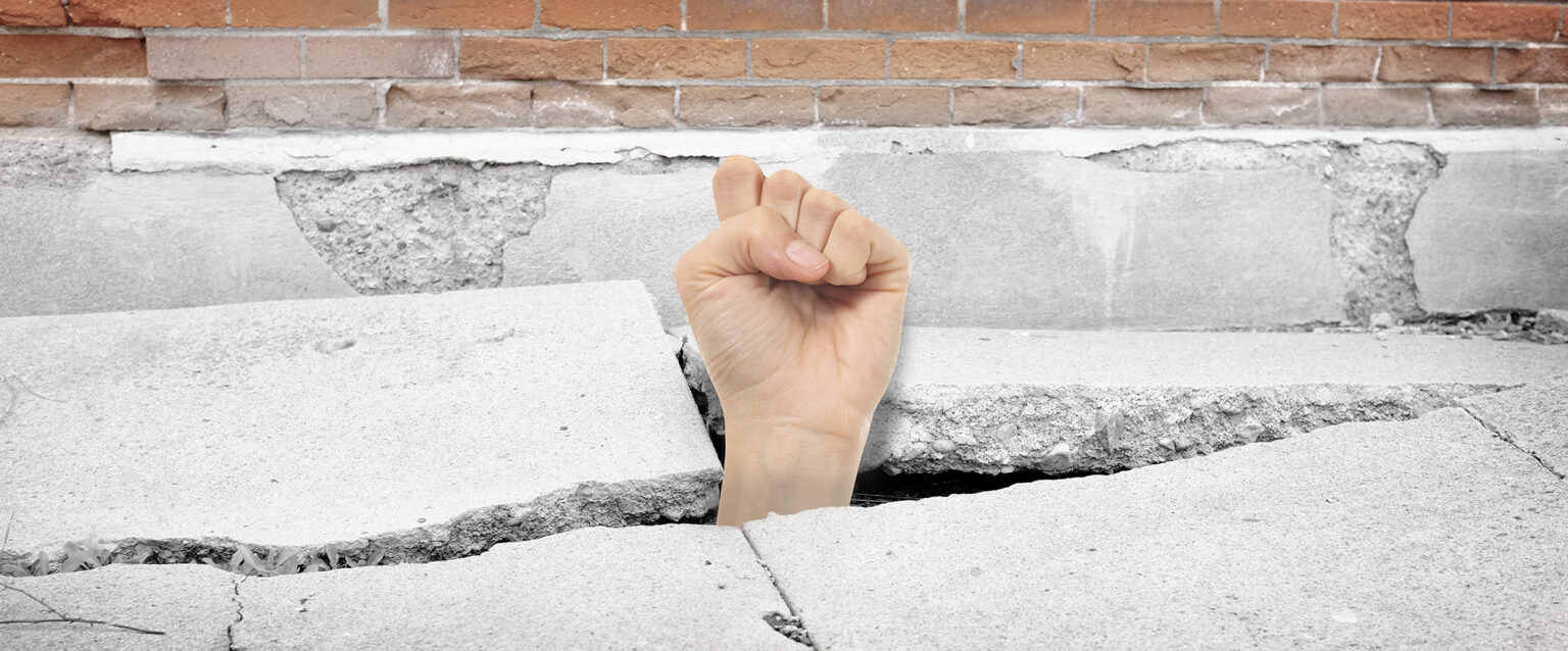 Cracked sidewalk concrete with a woman's fist breaking through from below.