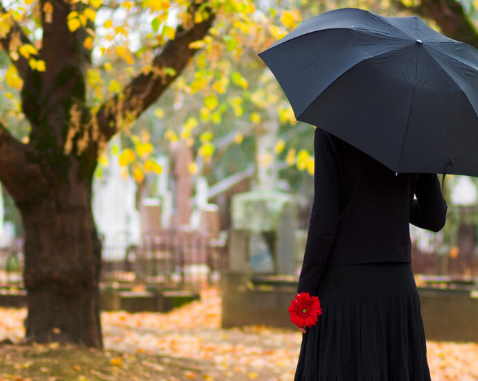 A woman in mourning, in a black coat and dress, with a black umbrella and a single red flower in her hand, faces a cemetery in Fall.
