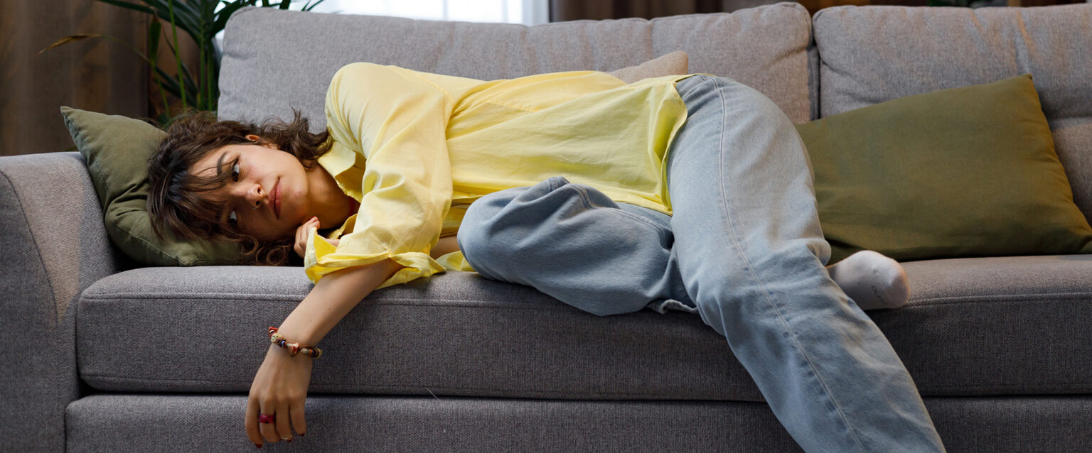 A girl in jeans and a yellow button down blouse, feeling a lack of motivation, falls over to lie on a gray couch.