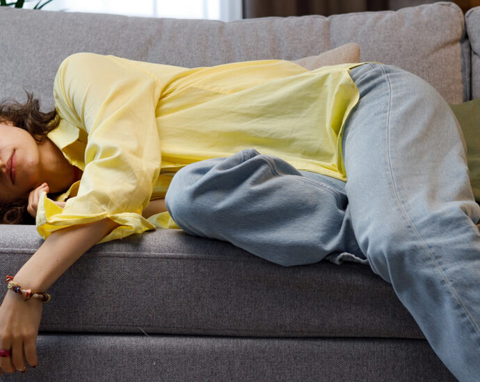 A girl in jeans and a yellow button down blouse, feeling a lack of motivation, falls over to lie on a gray couch.