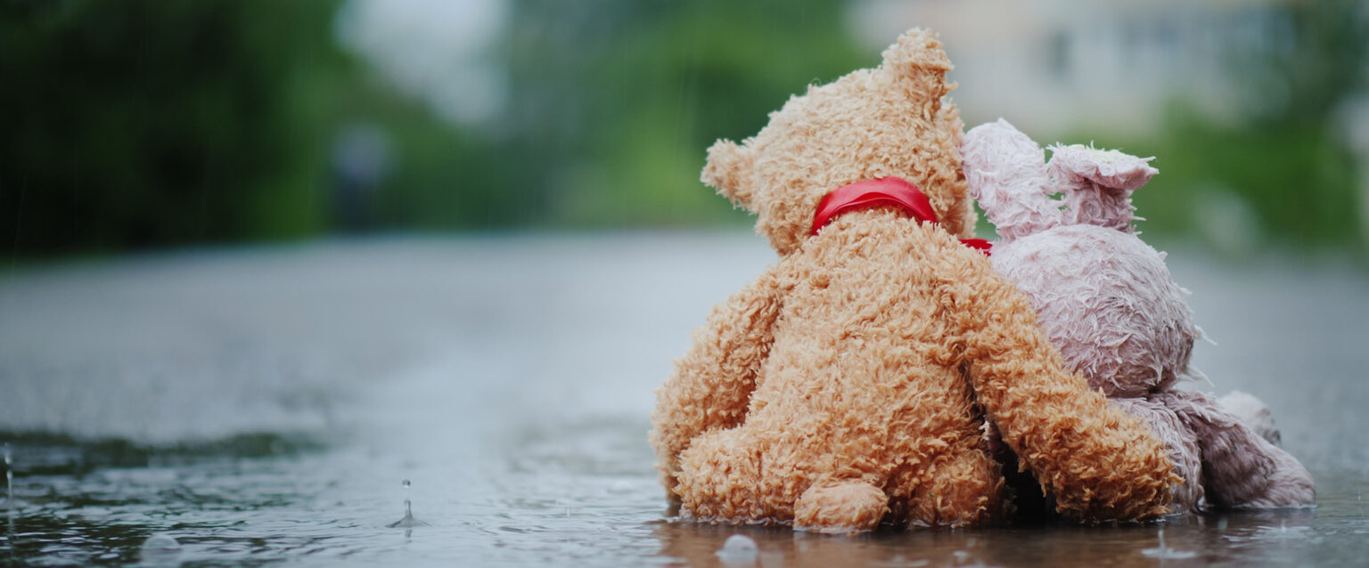 A pink, stuffed bunny, with one ear bent backward at the halfway point, and a brown teddy bear, with a red ribbon around its neck, sit side by side on a wet road in pouring rain. The bear's arm is around the bunny's back, and its head is tilted toward the bunny, as if showing compassion and care.