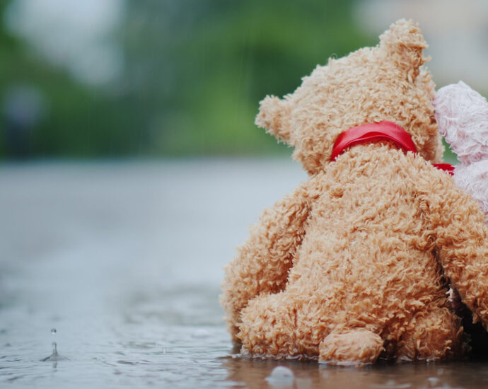 A pink, stuffed bunny, with one ear bent backward at the halfway point, and a brown teddy bear, with a red ribbon around its neck, sit side by side on a wet road in pouring rain. The bear's arm is around the bunny's back, and its head is tilted toward the bunny, as if showing compassion and care.