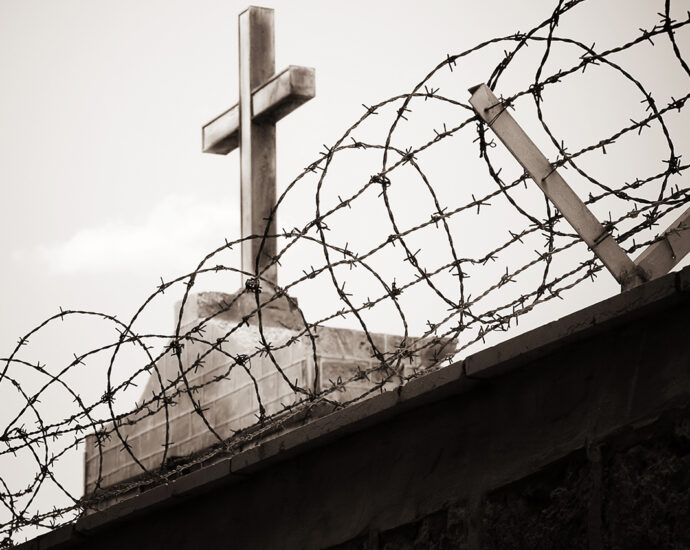The cross atop a church is visible as it rises above a dark, block wall with barbed wire across the top, in the foreground.