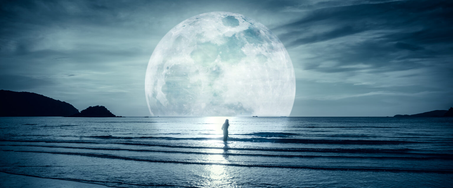 A photograph with cool tones, in which a woman stands in the water of a gulf as small waves roll into shore. The sky is cloudy and the moon is enlarged to emphasize the calming mood contained in the image.