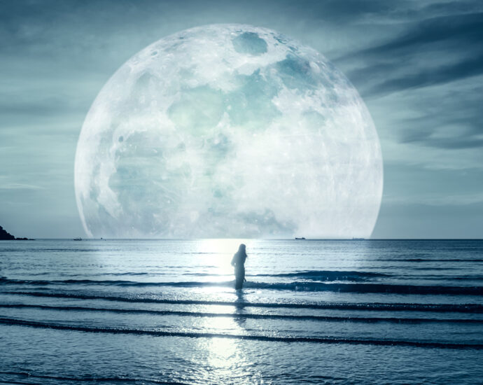 A photograph with cool tones, in which a woman stands in the water of a gulf as small waves roll into shore. The sky is cloudy and the moon is enlarged to emphasize the calming mood contained in the image.